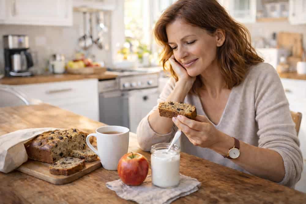 Femme de 40 ans prenant un petit-déjeuner équilibré avec banana bread, yaourt et thé dans une cuisine lumineuse